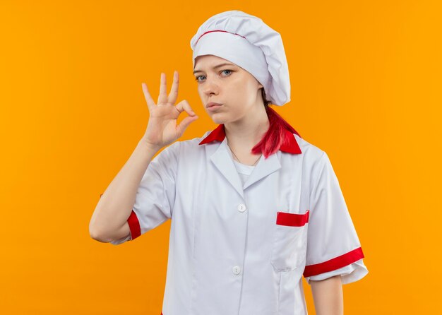 Joven chef mujer rubia segura en uniforme de chef gestos ok signo de mano y parece aislado en la pared naranja