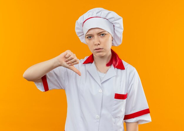 Joven chef mujer rubia segura en uniforme de chef pulgares hacia abajo y parece aislado en la pared naranja