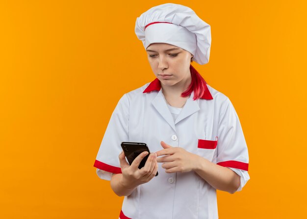 Joven chef mujer rubia segura en uniforme de chef sostiene y mira el teléfono aislado en la pared naranja