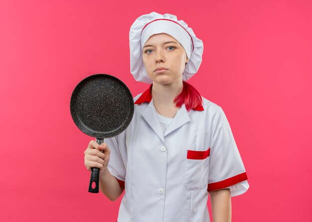 Joven chef mujer rubia segura en uniforme de chef sostiene una sartén y parece aislada en la pared rosa