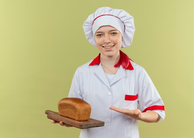 Joven chef mujer rubia sonriente en uniforme de chef sostiene y apunta al pan en la tabla de cortar aislada en la pared verde