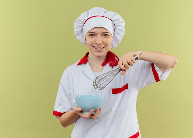 Joven chef mujer rubia sonriente en uniforme de chef sostiene tazón y batidor aislado en la pared verde