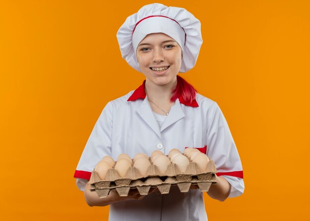 Joven chef mujer rubia sonriente en uniforme de chef tiene lote de huevos aislado en la pared naranja