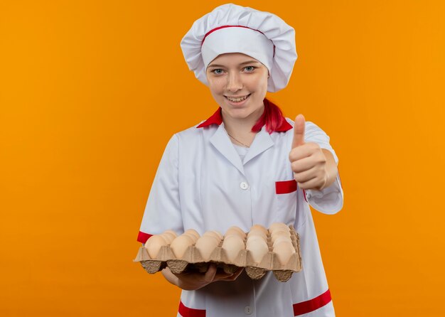 Joven chef mujer rubia sonriente en uniforme de chef tiene lote de huevos y pulgares arriba aislados en la pared naranja