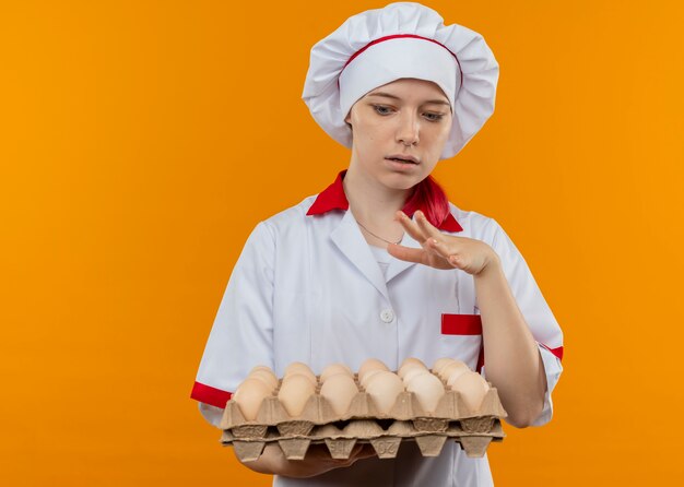 Joven chef mujer rubia sorprendida en uniforme de chef sostiene y mira el lote de huevos aislados en la pared naranja