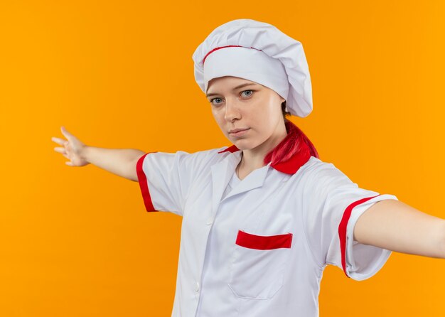 Joven chef mujer rubia en uniforme de chef se encuentra con los brazos abiertos aislados en la pared naranja