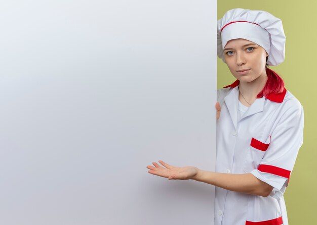 Joven chef rubia complacida en uniforme de chef está detrás y apunta a la pared blanca aislada en la pared verde