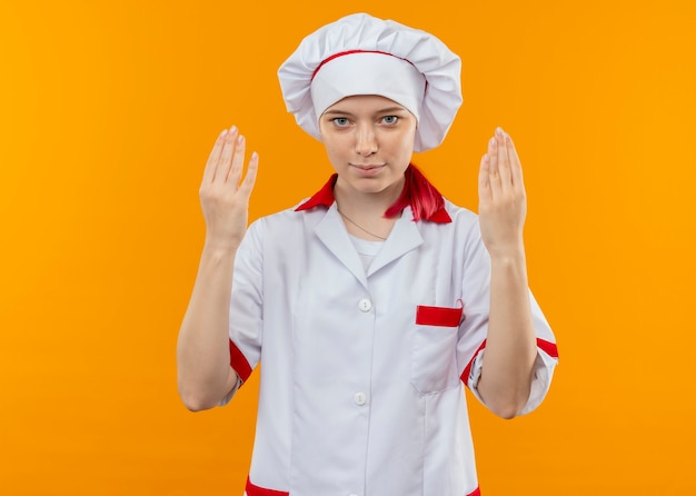 Joven chef rubia complacida en uniforme de chef levanta las manos aisladas en la pared naranja