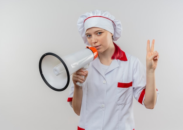 Joven chef rubia complacida en uniforme de chef sostiene altavoz y gestos signo de mano de victoria aislado en la pared blanca