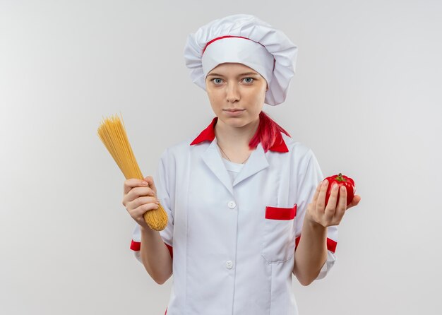 Joven chef rubia complacida en uniforme de chef sostiene un montón de espaguetis y pimiento rojo aislado en la pared blanca