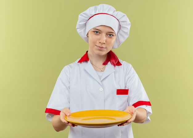 Joven chef rubia complacida en uniforme de chef sostiene la placa aislada en la pared verde