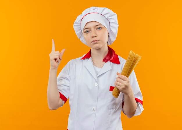 Joven chef rubia confiada en uniforme de chef sostiene un montón de espaguetis y apunta hacia arriba aislado en la pared naranja