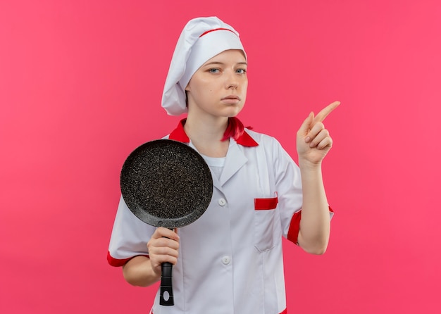 Joven chef rubia confiada en uniforme de chef sostiene una sartén y apunta hacia el lado aislado en la pared rosa