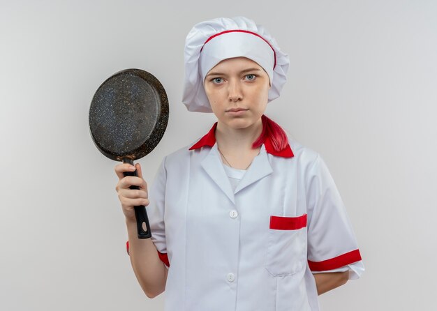 Joven chef rubia confiada en uniforme de chef sostiene una sartén y mantiene la mano detrás aislada en la pared blanca