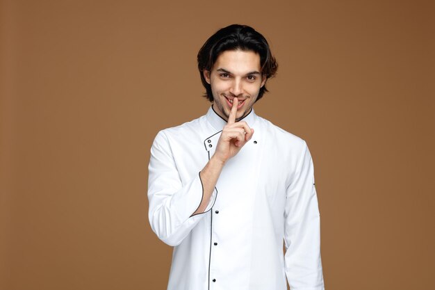 Un joven chef sonriente con uniforme mirando a la cámara mostrando un gesto de silencio aislado de fondo marrón