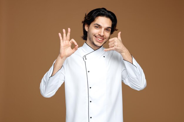 joven chef sonriente con uniforme mirando a la cámara mostrando el signo de ok y el gesto de llamada aislado en el fondo marrón