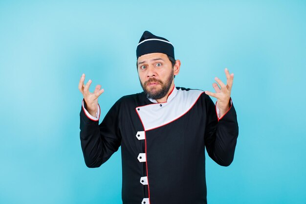 El joven chef sorprendido está mirando a la cámara levantando las manos sobre fondo azul.