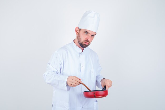 Joven chef en uniforme blanco mezclando comida con cuchara de madera y mirando pensativo, vista frontal.