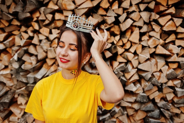 Joven chica divertida con maquillaje brillante como ropa de princesa de cuento de hadas en camisa amarilla y corona sobre fondo de madera