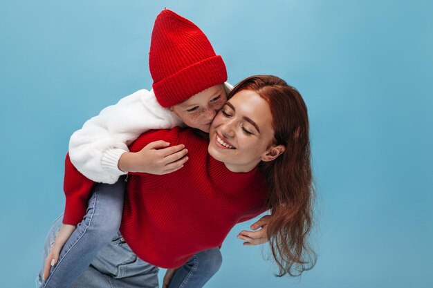 Joven chica elegante con pecas en traje blanco y taza roja besando a su hermana sonriente en suéter brillante sobre fondo azul.