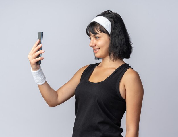 Joven chica fitness con diadema sosteniendo un teléfono inteligente mirándolo con una sonrisa en la cara feliz de pie sobre fondo blanco