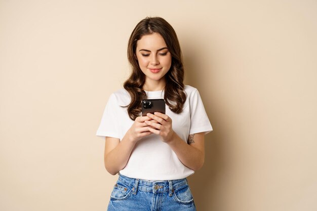 Joven chica moderna chateando en la aplicación usando la aplicación de teléfono inteligente y sonriendo de pie sobre un fondo beige