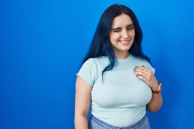 Joven chica moderna con el pelo azul sobre fondo azul guiñando un ojo mirando a la cámara con expresión sexy, cara alegre y feliz.