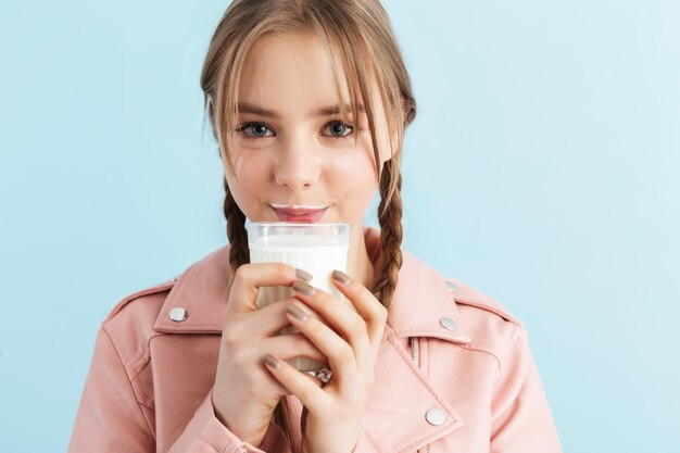 Joven chica muy sonriente con dos trenzas en chaqueta de cuero rosa con bigote de leche sosteniendo un vaso en la mano mientras mira soñadoramente en cámara sobre fondo azul