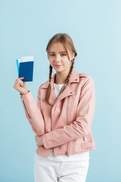 Joven chica muy sonriente con dos trenzas en chaqueta de cuero rosa y camiseta blanca con pasaporte con boletos en la mano mientras mira felizmente en cámara sobre fondo azul