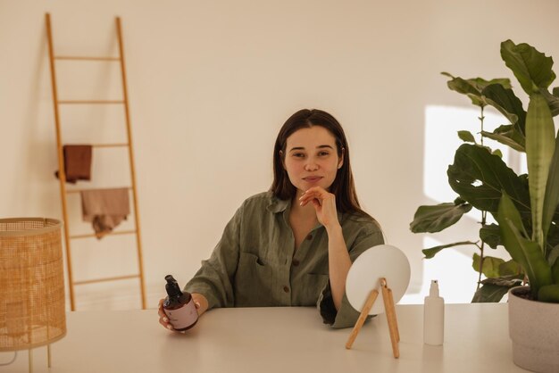 Joven chica de piel clara sostiene una botella grande de crema facial mientras mira a la cámara Morena de pelo largo con camisa se sienta a la mesa con espejo Señora posando en el concepto de casa