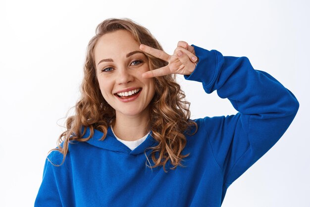 Joven chica rubia positiva muestra el signo de la paz haciendo un gesto de vsign y sonriendo feliz expresando una actitud optimista de pie sobre fondo blanco