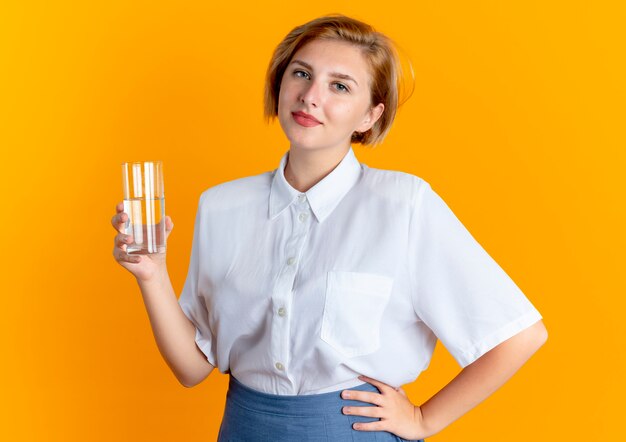 Joven chica rusa rubia complacida sostiene un vaso de agua aislado sobre fondo naranja con espacio de copia