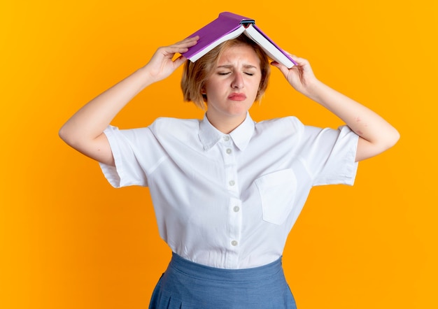 Joven chica rusa rubia molesta sostiene un libro sobre la cabeza aislado sobre fondo naranja con espacio de copia