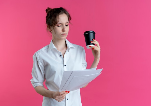 Joven chica rusa rubia segura mira hojas de papel sosteniendo la taza de café aislada en el espacio rosa con espacio de copia