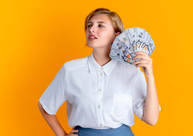 Joven chica rusa rubia segura tiene dinero mirando al lado aislado sobre fondo naranja con espacio de copia