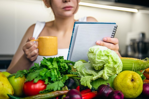 Una joven en la cocina con un cuaderno entre verduras.