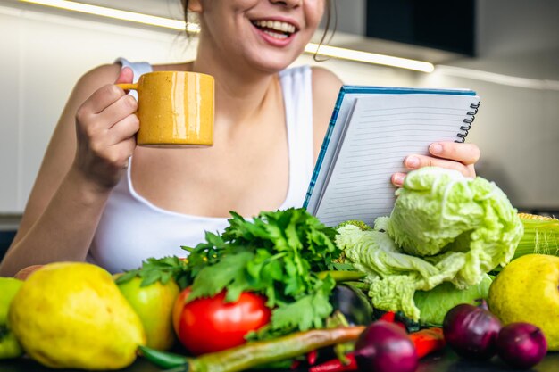 Una joven en la cocina con un cuaderno entre verduras.