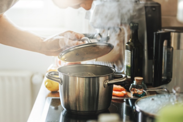 Joven cocinando alimentos frescos en casa y abriendo la tapa de la olla humeante.