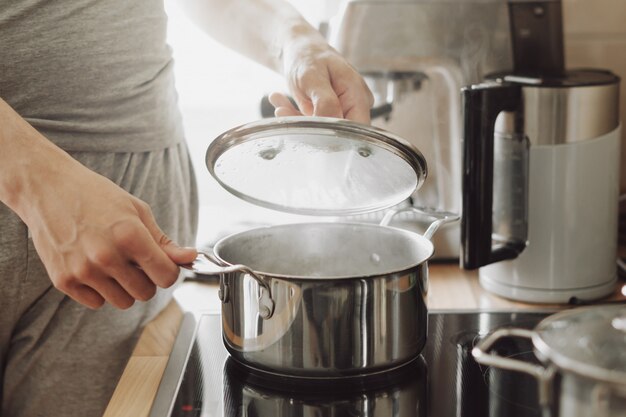 Joven cocinando alimentos frescos en casa y abriendo la tapa de la olla humeante.