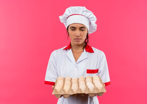 Joven cocinera aburrida vistiendo uniforme de chef sosteniendo un lote de huevos en una pared rosa aislada con espacio de copia