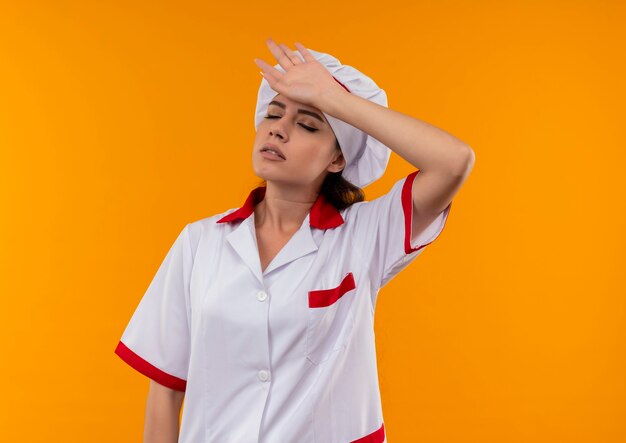 Joven cocinera caucásica cansada en uniforme de chef pone la mano en la cabeza con los ojos cerrados aislado en la pared naranja con espacio de copia