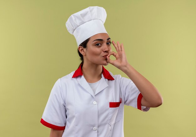 Joven cocinera caucásica complacida en chef uniforme gestos signo de mano delicioso sabroso aislado en la pared verde con espacio de copia