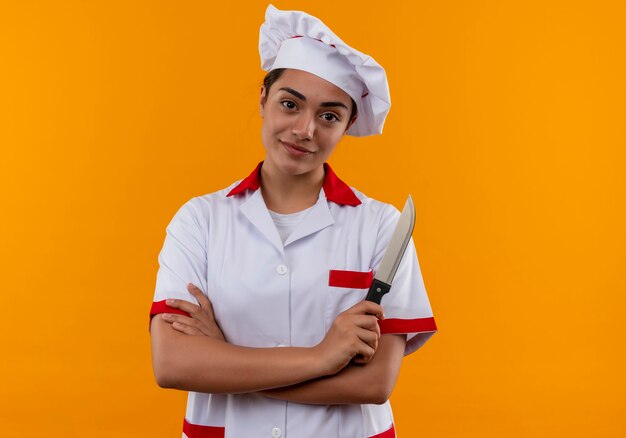 Joven cocinera caucásica complacida en uniforme de chef cruza los brazos y sostiene un cuchillo aislado en la pared naranja con espacio de copia