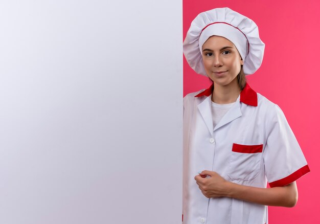 Joven cocinera caucásica complacida en uniforme de chef está detrás de la pared blanca en rosa con espacio de copia