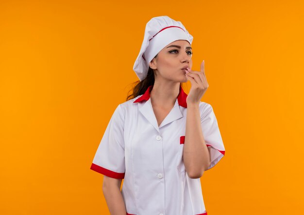 Joven cocinera caucásica complacida en uniforme de chef gestos signo de mano delicioso delicioso aislado en pared naranja con espacio de copia