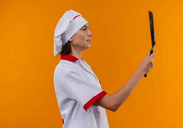 Joven cocinera caucásica complacida en uniforme de chef de pie lateralmente sostiene y mira la sartén aislada en el espacio naranja con espacio de copia