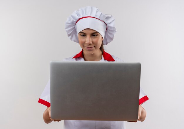 Joven cocinera caucásica complacida en uniforme de chef sostiene y mira portátil en blanco con espacio de copia