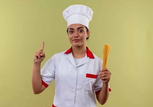 Joven cocinera caucásica complacida en uniforme de chef sostiene un montón de espaguetis y apunta hacia arriba aislado en la pared verde con espacio de copia