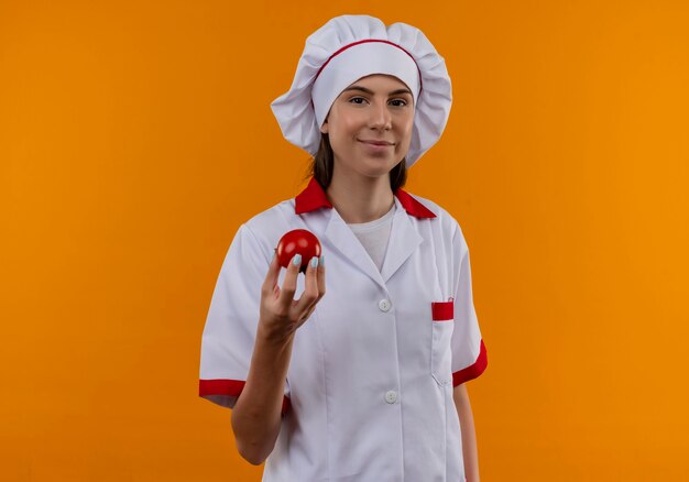 Joven cocinera caucásica complacida en uniforme de chef sostiene tomate mirando a cámara en naranja con espacio de copia