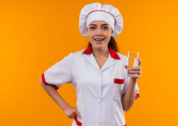 Joven cocinera caucásica complacida en uniforme de chef sostiene un vaso de agua aislado en la pared naranja con espacio de copia